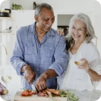 Couple stands in kitchen together, while man chops vegetables and woman smiles with a wine glass in her hand.